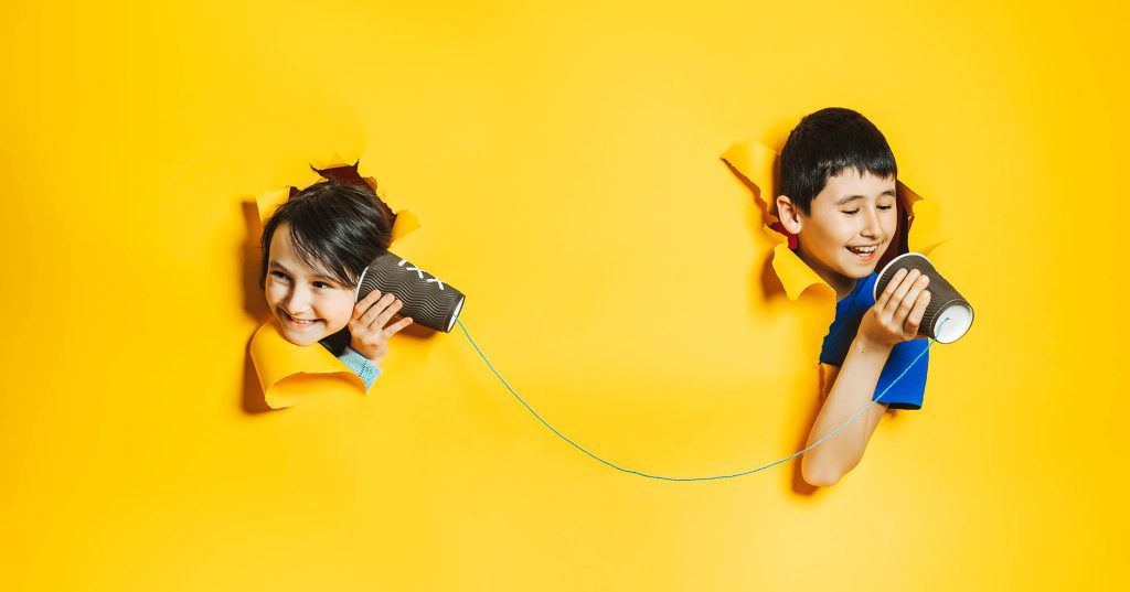 Kaksi hymyilevää lasta kurkistelee keltaisessa pahviseinässä olevista rei'istä, he keskustelevat toistensa kanssa mukeista ja langasta tehdyn puhelimen välityksellä.