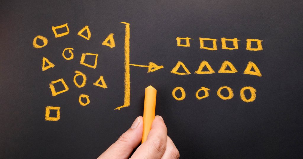 Liitutaululle on vasemmalle piirrretty eri muotoisia kuvioita, ne ovat sekaisin. Oikealle ne on järjestetty selkeille riveille kuvioittain. Taulun edessä on käsi liidun kanssa.