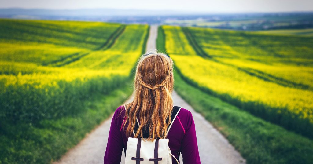 Ylä-asteikäinen tyttö reppu selässä pitkän tien päässä, katsoo yksin eteenpäin horisonttiin. Ympärillä keltaisia peltoja.